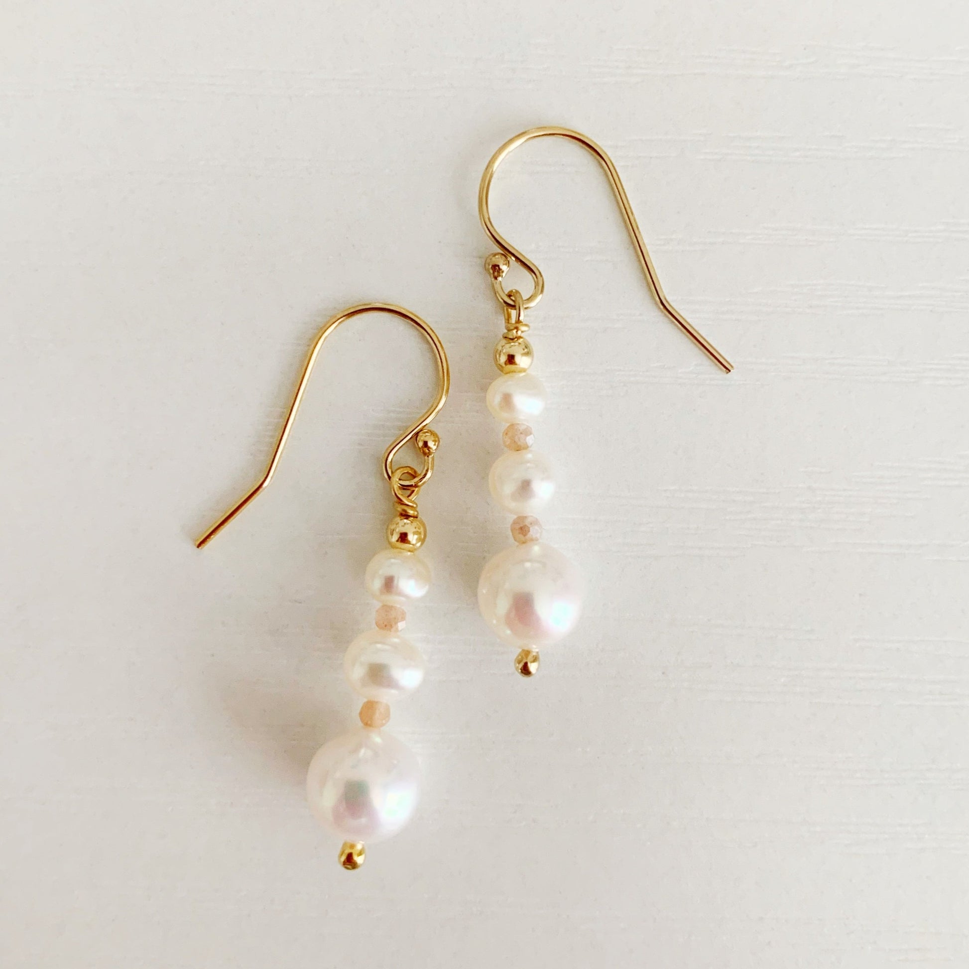 Barrington earrings by mermaids and madeleines feature freshwater pearls in a linear taper with peach moonstone accents and 14k gold filled findings. this pair is photographed flat on a white surface