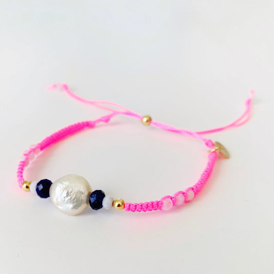 The bristol macramé bracelet in color shrimp on the barbi is a neon pink bracelet with a pearl and semiprecious beads at the center and a 14k gold filled slide clasp at the back. this bracelet is photographed on a white surface