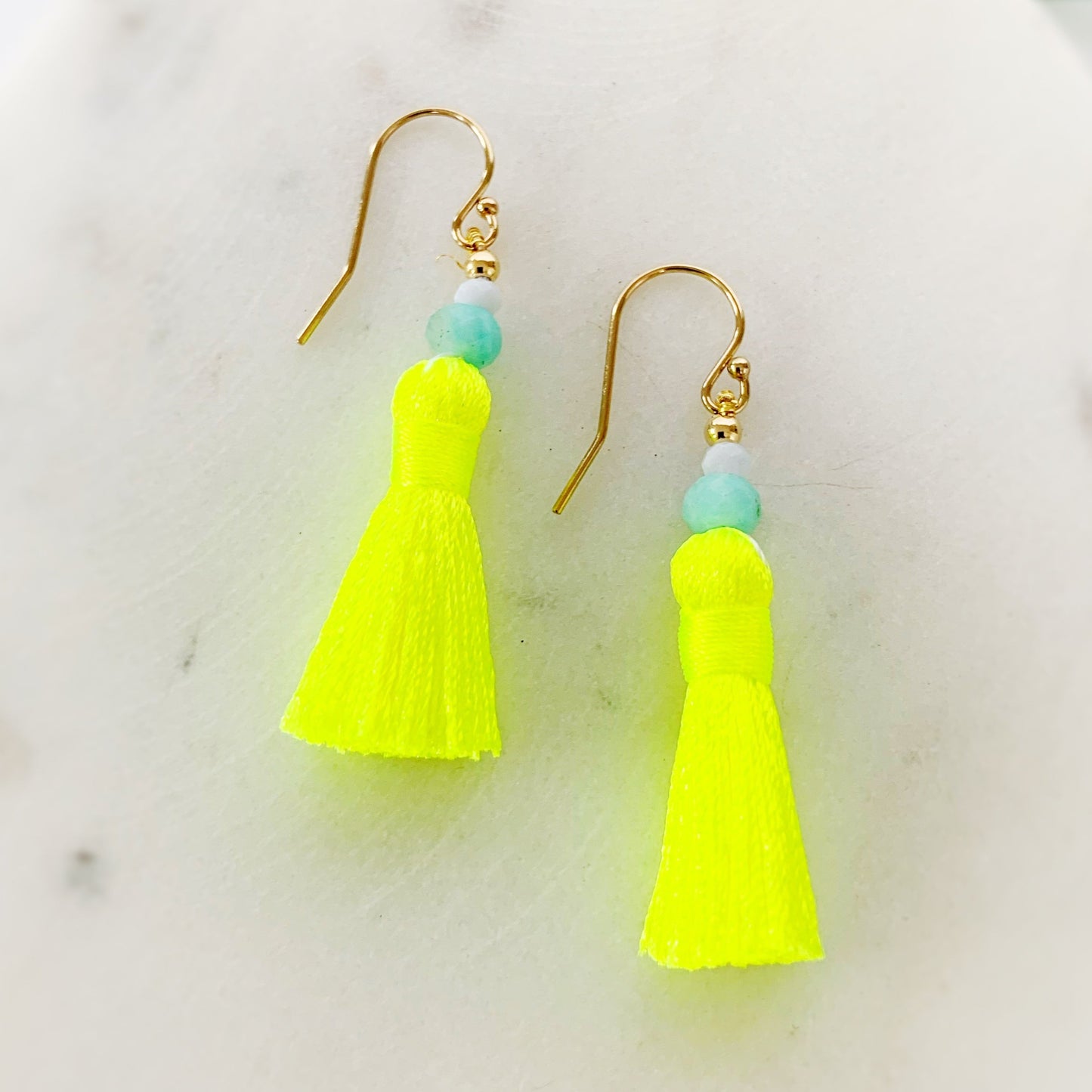 a close up view of the captiva tassel earrings from the mermaids and madeleines sunshine collection. The earrings have bright neon yellow tassels and amazonite and blue lace agate beads with 14k gold filled earring findings. This pair is photographed on a white lightly marbled surface