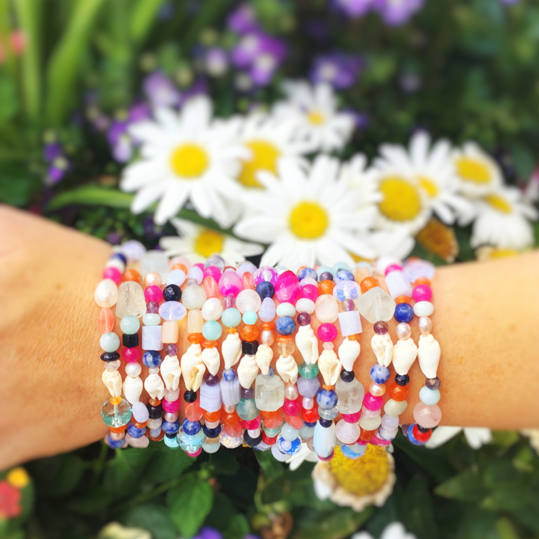 a wrist stacked full of treasure bracelets and photographed over a planter of daisies. The treasure bracelets are created with multicolor semiprecious beads, freshwater pearl and shell on a stretchy bracelet