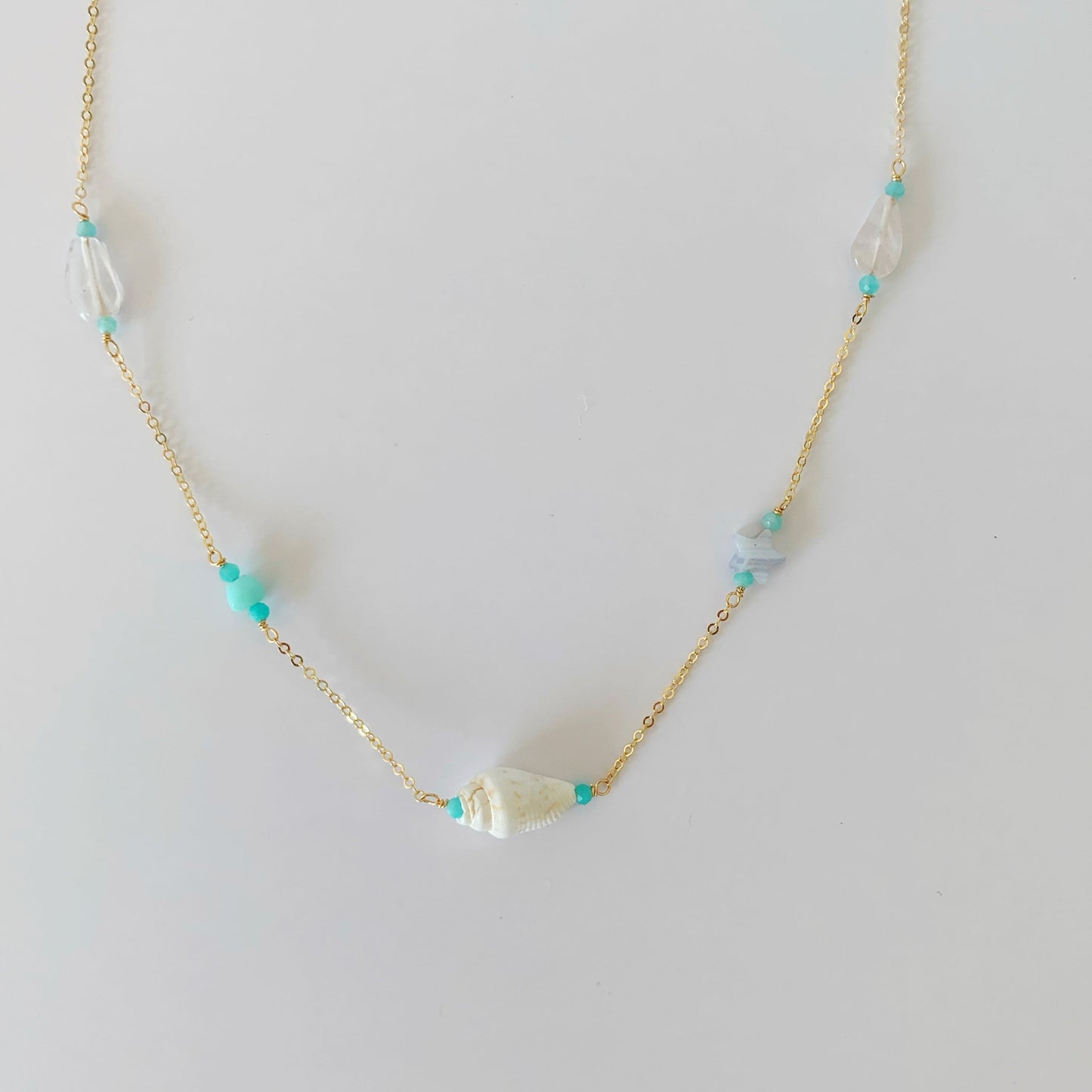The island hopper necklace by mermaids and madeleines is a station style necklace with semiprecious beads and a shell at the center on 14k gold filled chain. this necklace is photographed on a white surface