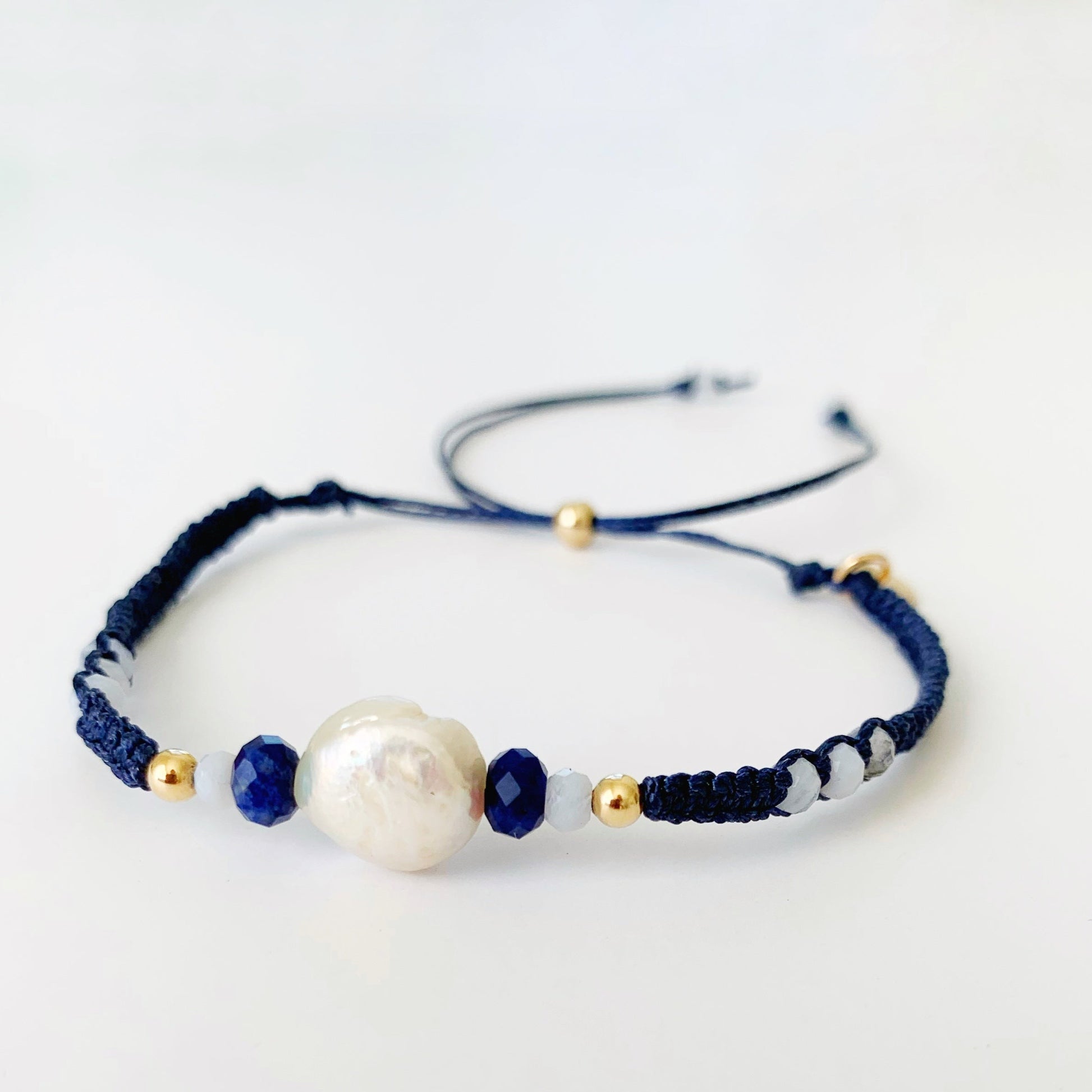 The bristol bracelet in knotical navy is a macrame bracelet in navy blue cord with a freshwater coin pearl and semiprecious beads at the center with a 14k gold filled slide bead clasp. this bracelet is photographed on a white surface