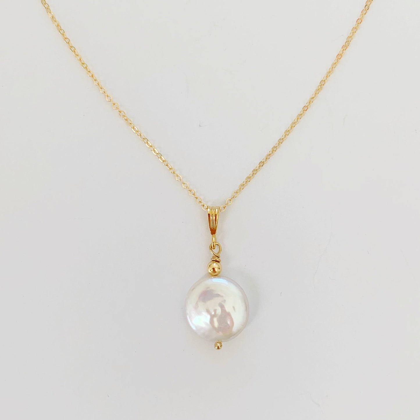 A close up view of the newport gala necklace from mermaids and madeleines. this necklace is a large freshwater coin pearl pendant with 14k gold filled findings and chain and its photographed on a white surface