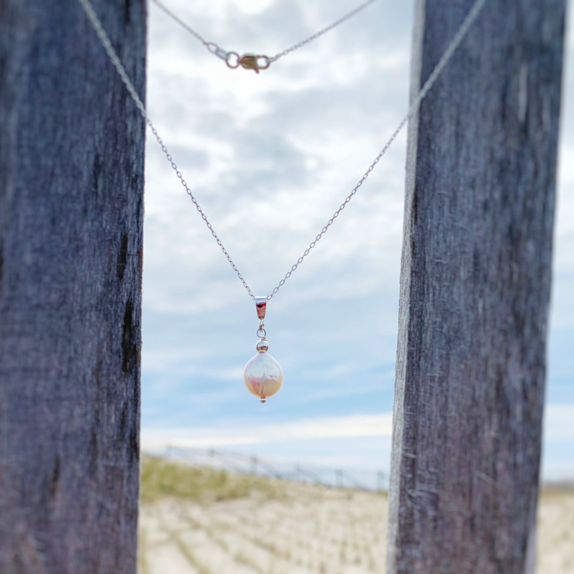  the Newport pendant necklace from the mermaids and madeleines tide pool collection.The necklace is created with a freshwater white iridescent coin pearl suspended from a sterling silver bail and chain. It's photographed here hanging on a beach fence with blurred fence posts and beach in the background
