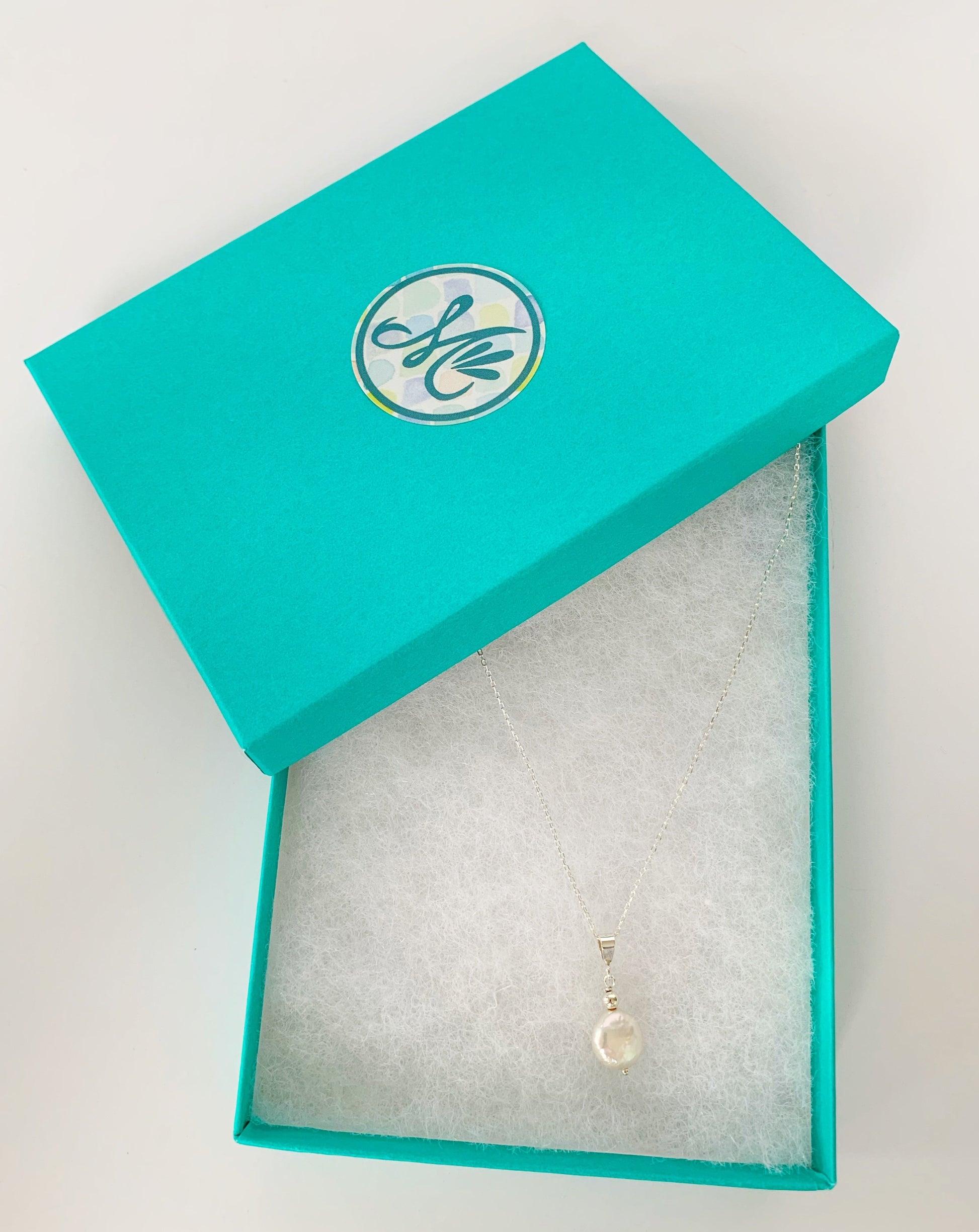 The Newport pendant necklace pictured in a cotton lined teal gift box with a mermaids and madeleines sticker on the lid. The necklace is created with a white freshwater coin pearl suspended from a sterling silver bail and chain. The item is photographed on a white background