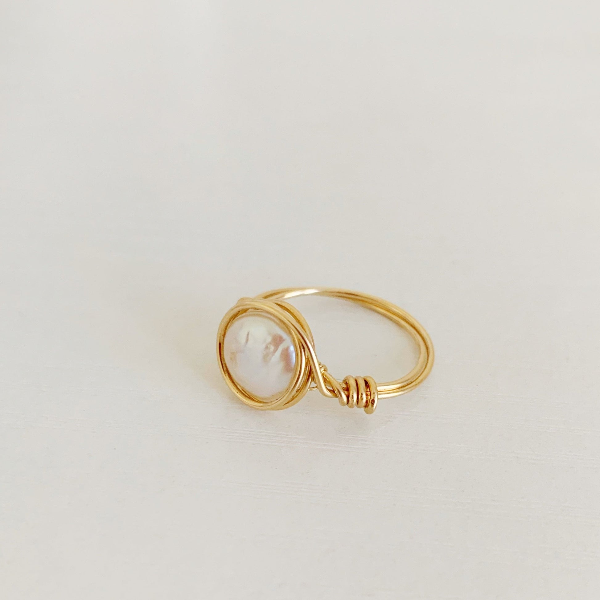 Newport Pearl Ring in 14k gold filled by mermaids and madeleines is a wire wrapped ring that features a freshwater white coin pearl wire wrapped in 14k gold filled wire creating an artful and simple ring. this one is photographed at an angle on a white surface