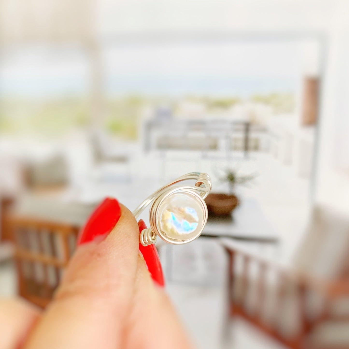 the newport pearl ring in sterling silver by mermaids and madeleines features a freshwater white coin pearl in a wire wrap ring with sterling silver wire. the design is simple and artful. this ring is photographed held between fingers in front of a blurred living room scene