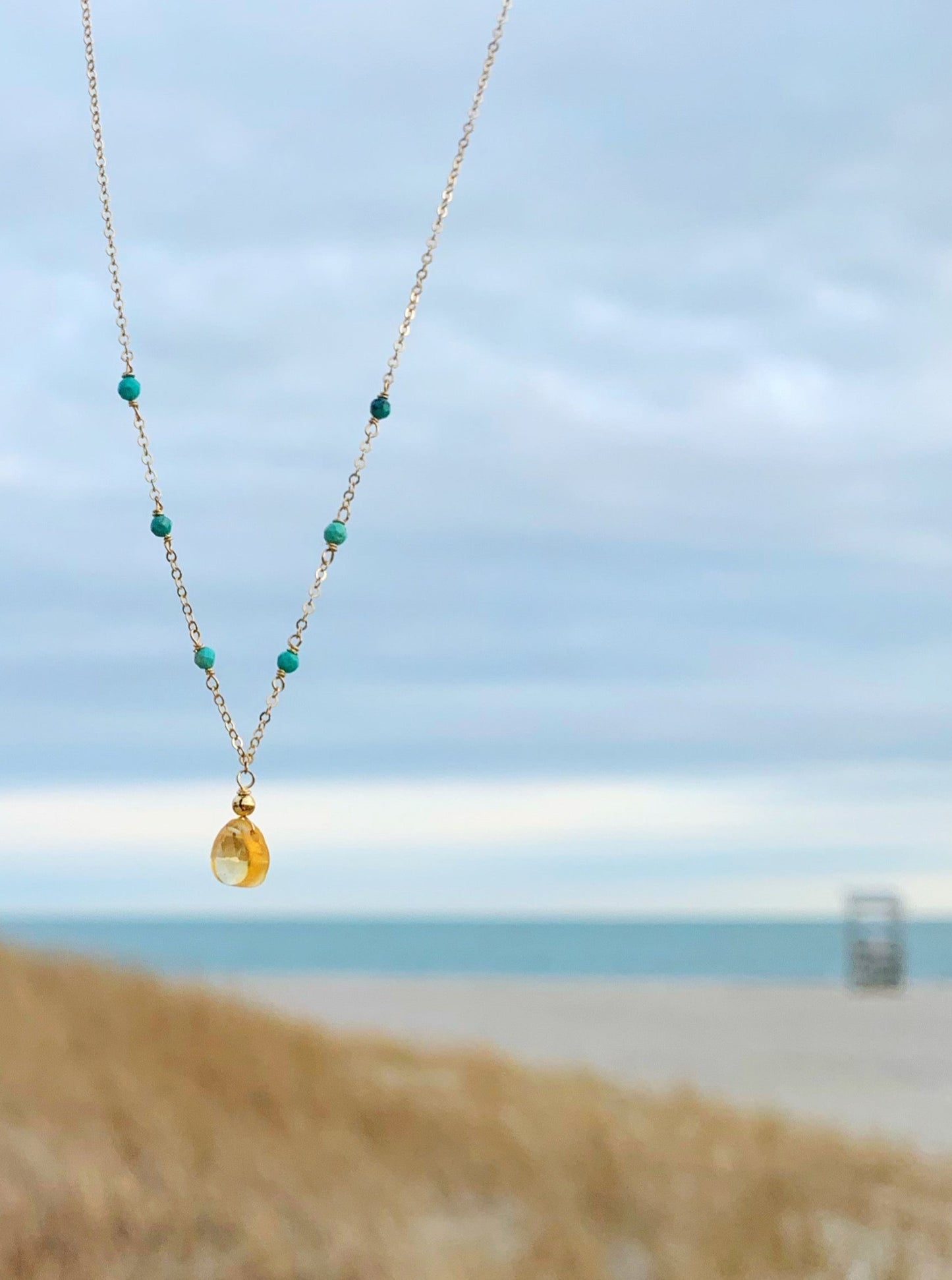 Ray of Sunshine necklace by mermaids and madeleines is created with a citrine drop at the center and natural turquoise beads on a 14k gold chain. this necklace is pictured in front of a beach scene