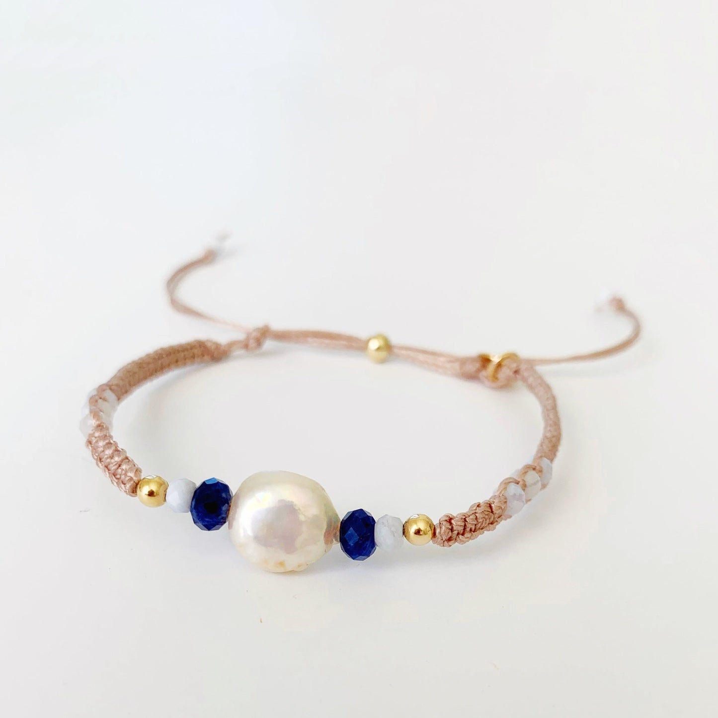 The bristol macrame bracelet in color iced coffee addict is a tan adjustable cord bracelet with freshwater coin pearl and semiprecious beads at the center and a 14k gold filled slide clasp in the back. this bracelet is photographed on a white surface