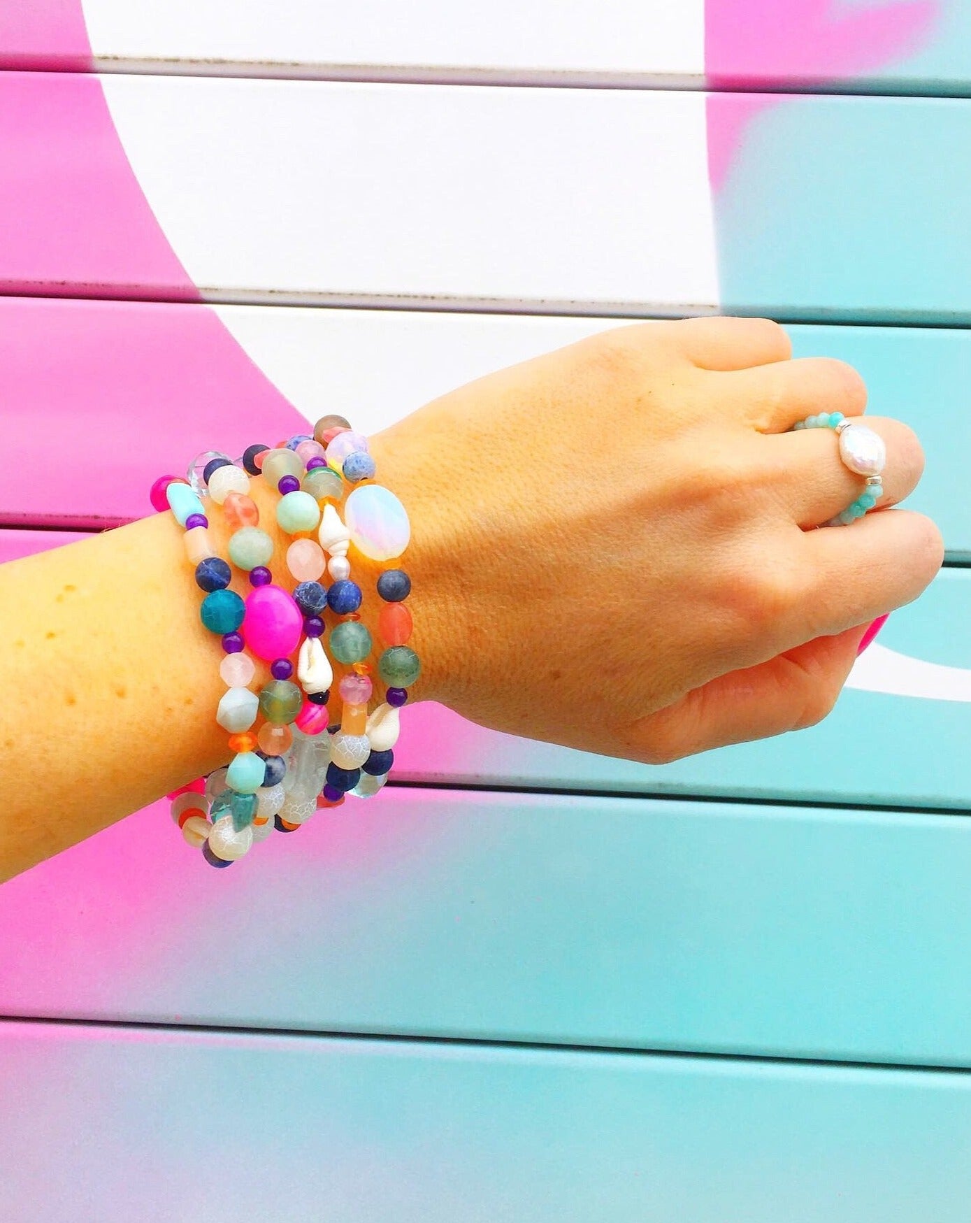 A picture of a wrist stacked with 5 treasure bracelets photographed in front of a teal and pink mural. The treasure bracelets are bright mulitcolor semiprecious beads with freshwater pearl and a beach shell on a stretchy bracelet