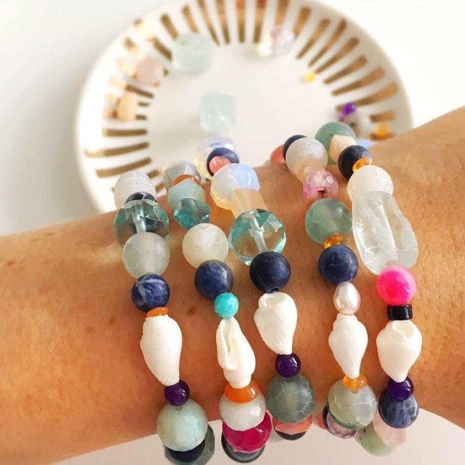 A wrist stacked with 5 treasure bracelets on it and in front of a white background and a small white and gold trinket dish. The treasure bracelets are created with various multicolor semiprecious beads, freshwater pearl and a shell on a stretchy bracelet