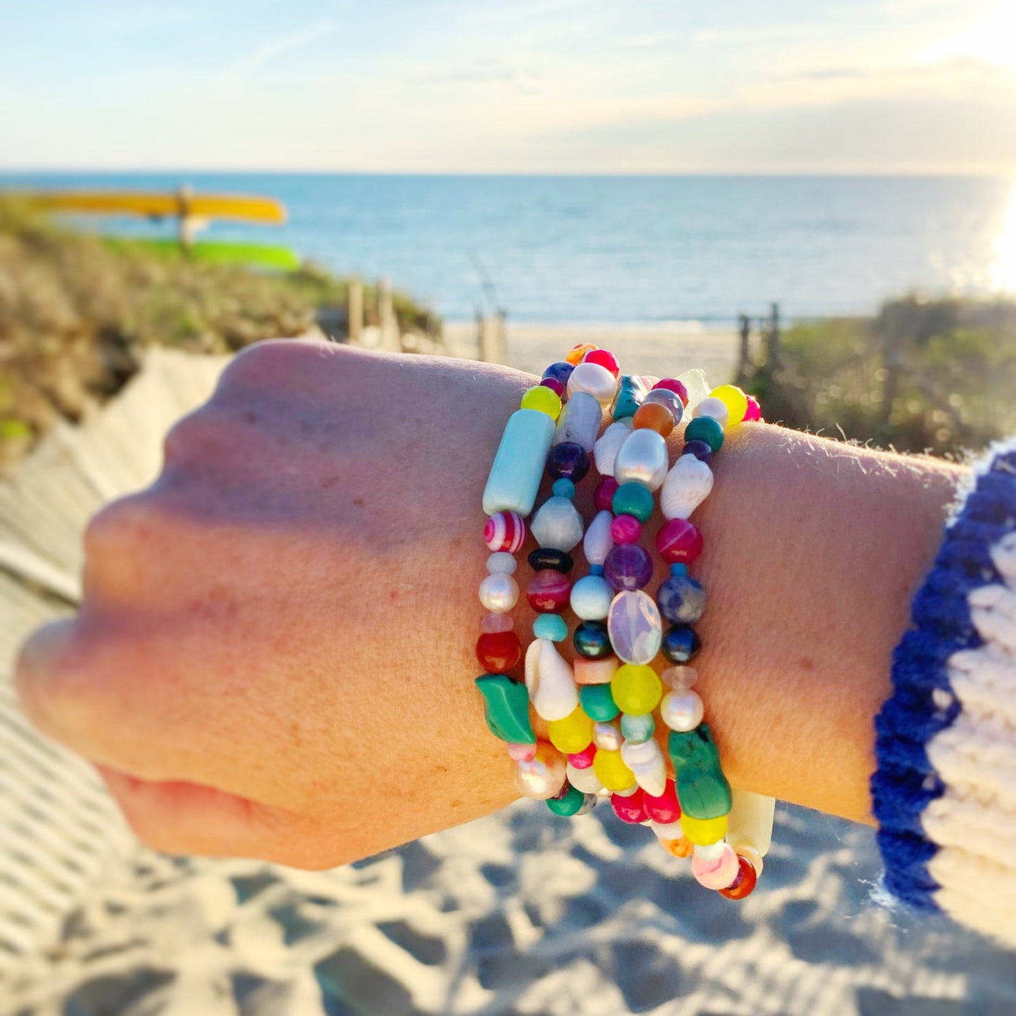 Treasure bracelets are a mix of bright, precious and semiprecious stones and freshwater pearls on a stretchy bracelet. Pictured is a wrist stacked with 5 treasure bracelets held up in front of a beach scene