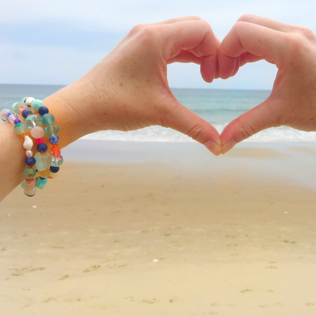 an image of 2 hands forming a heart shape at the beach. One wrist is shown stacked with 3 treasure bracelets on it. The treasure bracelets are created with multicolor semiprecious beads, freshwater pearls and shell on a stretchy bracelet