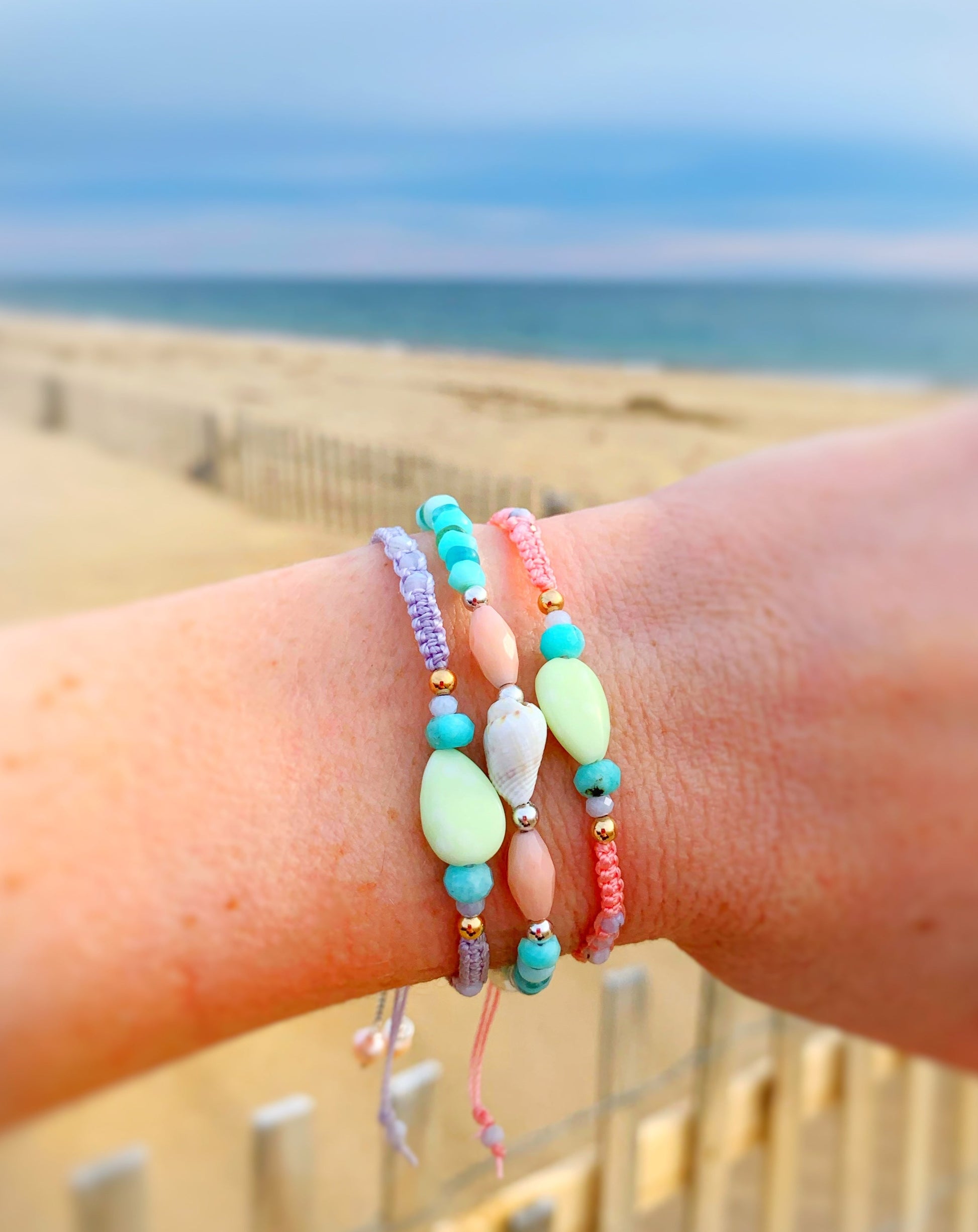 A picture of the Captiva tropical smoothie macrame bracelet which is peach color, on a wrist wearing 2 other bracelets captiva in coastal haze and the shellebration bracelet in pool party. Photographed with beach in the background
