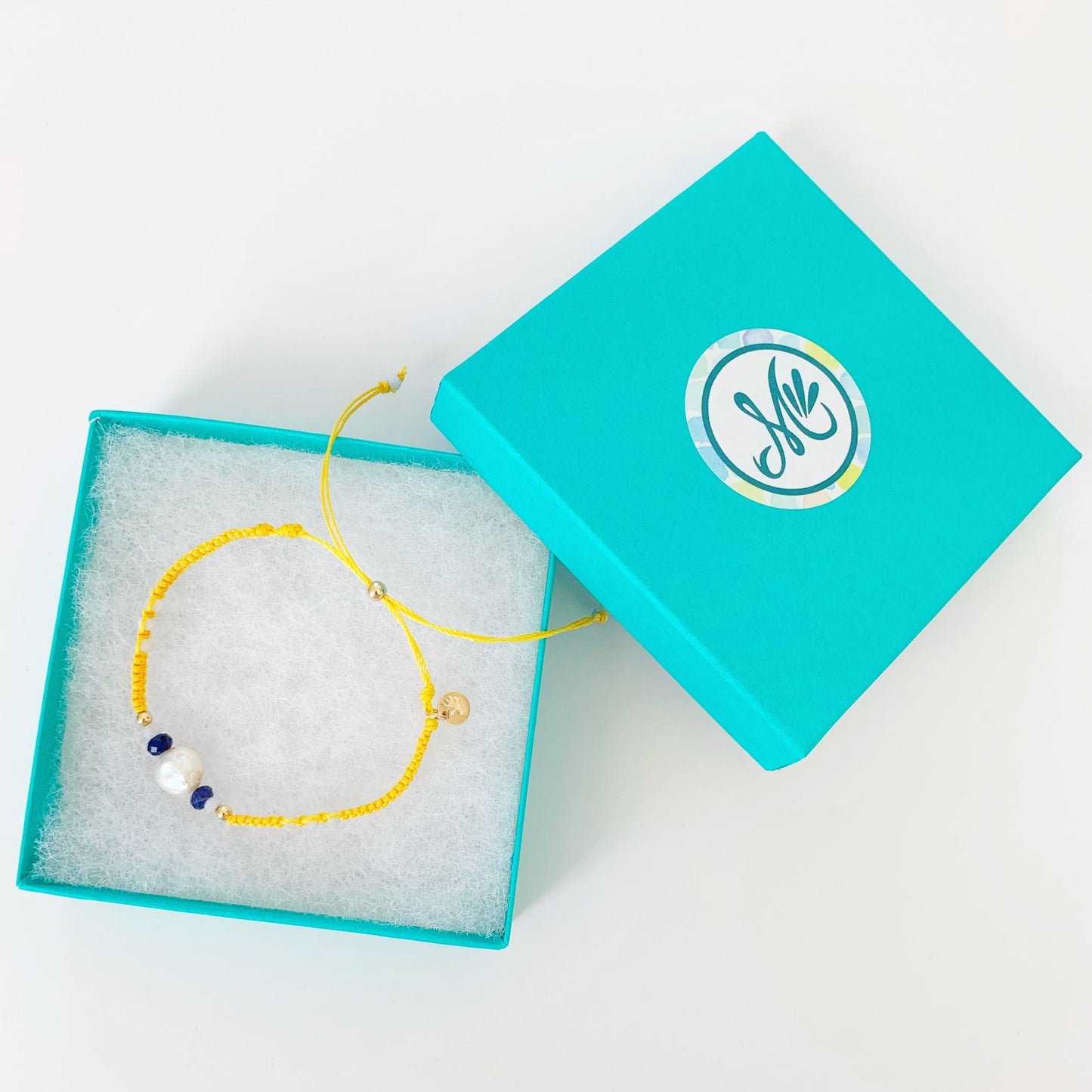 The yellow macrame bracelet is photographed in a teal mermaids and madeleines gift box on a white surface